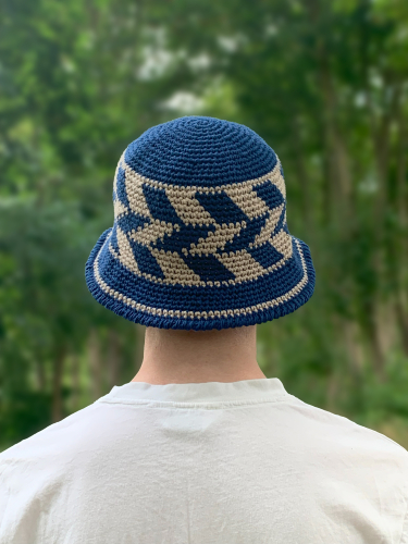 Mand i hvid T-shirt med bøllehat på med geometrisk mønster med skrå striber i mørkeblå og sand på siden af hatten. Manden står med ryggen til, og der er træer i baggrunden. 