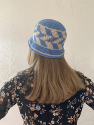 Kvinde, der står med ryggen til og har en bøllehat på med geometrisk mønster med skrå striber i mellemblå og sand. Kvinden har langt hår og en blomstret bluse på.