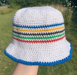 Hvid bøllehat med fem striber i blå, gul, sort, grøn og rød på siden af hatten og en blå kontrastkant forneden på en hånd. I baggrunden er der græs.