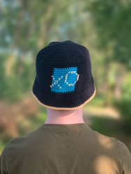 Sort bøllehat med turkis firkant på forsiden på hovedet af en mand i grøn T-shirt set bagfra. I firkanten er der broderet et X og et O med hvidt garn. Kanten på skyggen er hæklet i lys fersken. Træer i baggrunden.