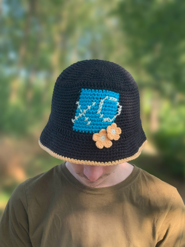 Sort bøllehat med turkis firkant på forsiden på hovedet af en mand i grøn T-shirt. I firkanten er der broderet et X og et O med hvidt garn, og i nederste højre hjørne af hatten er der to hæklede blomster i lys fersken. Kanten på skyggen er også hæklet i lys fersken. Træer i baggrunden.