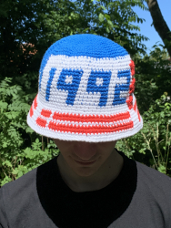 Opskrift på hæklet bøllehat som pdf fil. Hatten vises forfra, og den er blå på toppen, hvid foran med tallene 1992 i blå og på skyggen er der hæklet et dansk flag i rød og hvid. Ud for 2-tallet er der placeret tre røde stjerner.