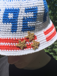 Opskrift på hæklet bøllehat som pdf fil. Billedet viser et nærbillede af skyggen på hatten, hvor tre hæklede guldstjerner er syet fast oven på to røde striber på den hvide skygge lige under 2-tallet på forsiden af hatten.